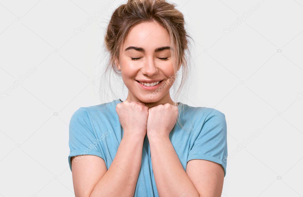 Grateful young woman smiling and keeps hands on chest with closed eyes, dressed in blue t-shirt, isolated over white background. People, emotions, feeling and body language concept