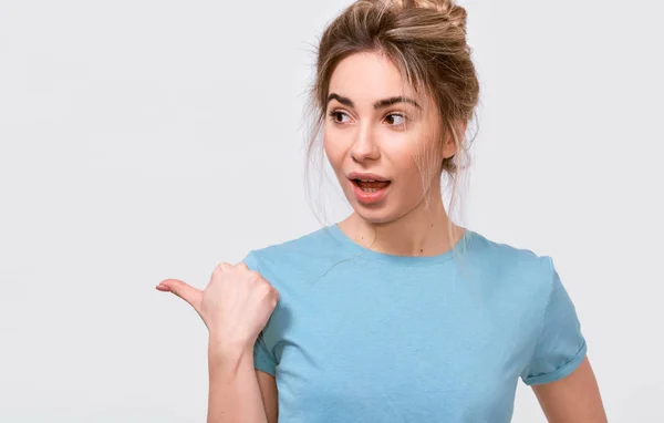 Jovem excitada europeia vestindo camiseta azul casual, indicando espaço em branco para texto publicitário, olhando para o lado, posando sobre fundo de estúdio cinza. Pessoas emoções — Fotografia de Stock