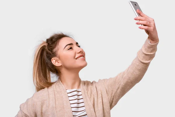 Side view studio shot of a pretty young European woman in casual clothes smiling to her boyfriend while speaking online. Lovely female taking a selfie isolated over white studio background. — Stock Photo, Image