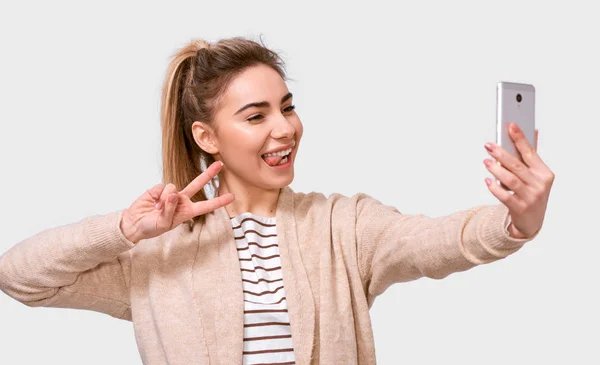 Studio bild av en lycklig ung europeisk kvinna i casual kläder visar tungan och fred tecken, tar en selfie isolerad över vit Studio bakgrund. — Stockfoto