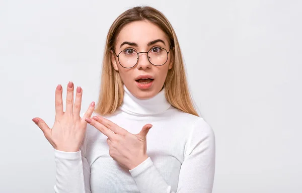 Portrait d'une belle jeune femme blonde excitée portant un chemisier blanc et des lunettes transparentes rondes à la mode, montrant une bague de fiançailles à son doigt, posant sur un fond de studio blanc . — Photo