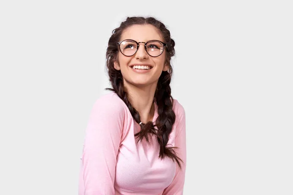 Чарівна молода жінка, яка посміхається широко, носить рожеву повсякденну блузку і круглі прозорі окуляри, дивлячись вгору, ізольовані на білому тлі. Щаслива молода жінка з зачіскою на сірій студійній стіні — стокове фото