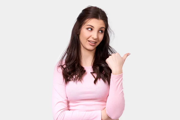 Horizontal studio image of beautiful European young woman wearing casual pink long sleeve blouse, indicating to blank copy space for advertising text, looking aside, posing over white background. — Stock Photo, Image