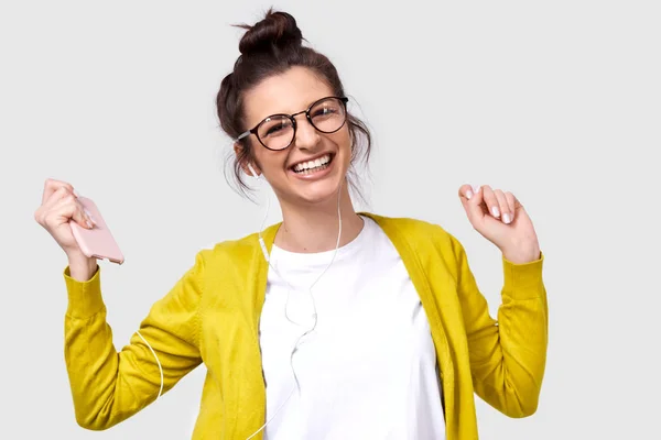 Jovem atraente e muito feliz sorrindo amplamente, usando o celular para ouvir a música. Feliz dança feminina e desfrutar das músicas favoritas via telefone inteligente em fones de ouvido. Emoções humanas reais — Fotografia de Stock