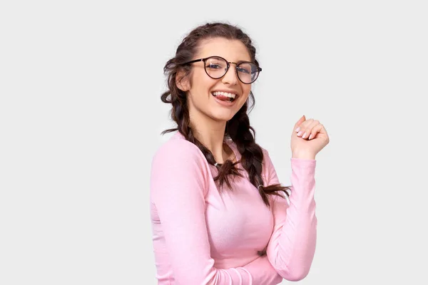 Позитивна мила молода жінка посміхається широко, носить рожеву повсякденну блузку та круглі прозорі окуляри, ізольовані на білому тлі. Щаслива молода жінка з зачіскою на сірій студійній стіні . — стокове фото