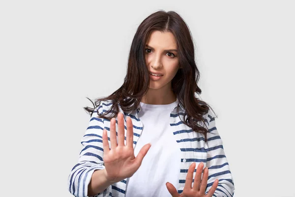 Fotografía de una joven morena haciendo un gesto de stop con las palmas de las manos. Retrato de mujer caucásica con camisa a rayas y camiseta blanca, haciendo gesto de rechazo, posando sobre fondo blanco. Negativo. — Foto de Stock