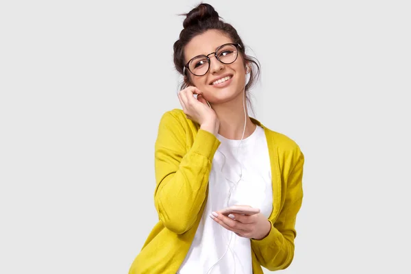 Studio poziome portret szczery uśmiechnięta kobieta w okrągłe okulary, dotykając słuchawek. Kryty strzał kaukaski młoda kobieta z błyszczącą skórą nosi żółte bluzki i biały t-shirt, cieszyć się muzyką. — Zdjęcie stockowe