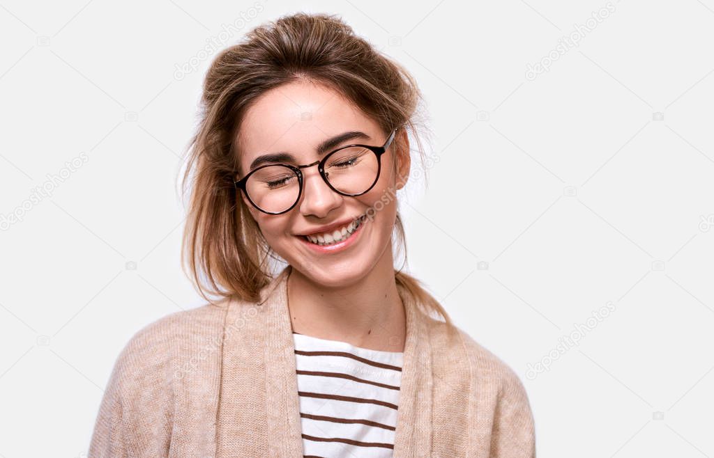 Horizontal close up portrait of dreamful positive female in casual outfit and eyewear, smiling with closed eyes, imagines something pleasant, posing over white background. People positive emotions