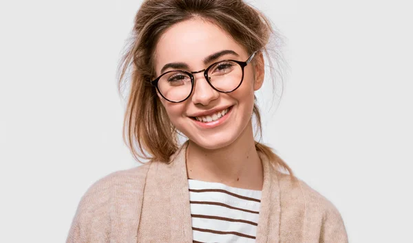 Close up retrato de bela fêmea positiva vestida com roupa casual, vestindo óculos redondos transparentes com sorriso agradável, olhando para a câmera e posando sobre a parede branca. Pessoas emoções — Fotografia de Stock