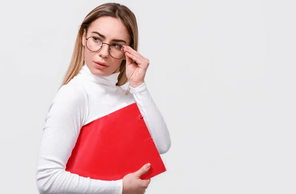 Jovem empresária séria vestindo blusa branca e óculos redondos da moda com pasta vermelha na mão, com espaço em branco para o seu texto. europeu feminino escritório trabalhador posando no branco estúdio parede . — Fotografia de Stock