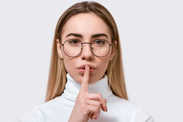 Close up portrait of beautiful young woman in round eyewear, holding her index finger to her lips to keep silence, on white wall. Pretty female wears spectacles, demonstrates secret sign, demands hush — Stock Photo, Image