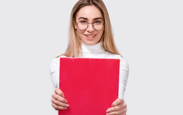 Retrato de estudante jovem bem sucedido vestindo blusa branca e óculos redondos transparentes com pasta vermelha nas mãos, sorrindo para a câmera. alegre feminino escritório trabalhador posando no branco parede . — Fotografia de Stock