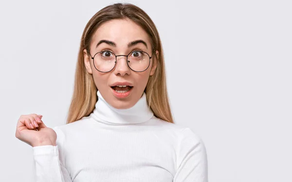 Amazed young woman wears eyewear and casual whote outfit, looking at camera, isolated on white studio background. Close up portrait of surprised female feels shocked. People emotions concept. — Stock Photo, Image