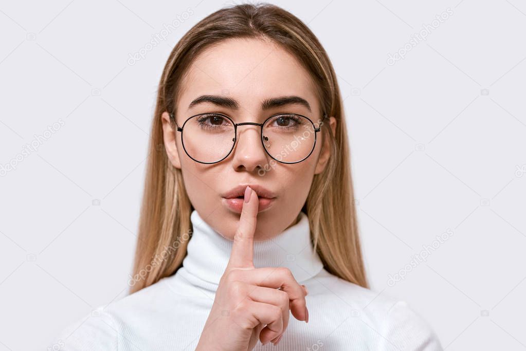 Close up portrait of beautiful young woman in round eyewear, holding her index finger to her lips to keep silence, on white wall. Pretty female wears spectacles, demonstrates secret sign, demands hush