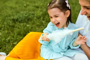 Mutlu çocuk gülümseyerek ve annesi ile oynarken, birlikte açık havada zaman zevk. Neşeli küçük kız annesiyle pamuk şeker yiyor, parkta yeşil çimenlerin üzerinde oturuyor. Anneler günü.