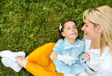 Parkta yeşil çimenlerin üzerinde oturan genç annesi ile pamuk şeker yiyen güzel kızı üst görünümü. Güzel kadın çocuğuna açık havada sarılıyor, birlikte vakit geçirdik. Anneler günü.