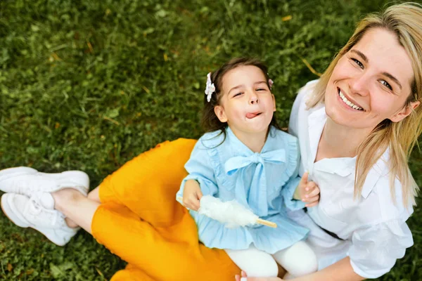 Szczęśliwa matka uśmiechając się i grając z dzieckiem, ciesząc się razem na zewnątrz. Radosna dziewczynka jedzenia Wata Cukrowa z mamą, siedząc na zielonej trawie w parku miejskim. Dzień matki. — Zdjęcie stockowe