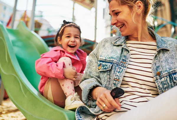 Image franche de jeune femme heureuse riant avec sa petite fille mignonne passant du temps ensemble à l'extérieur sur la glissière à l'aire de jeux. Mère avec son tout-petit fille se sentant heureux, s'aimant à l'extérieur . — Photo