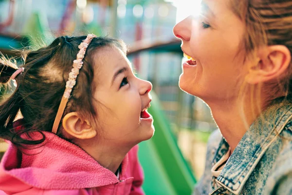 Zbliżenie portret szczęśliwej młodej kobiety uśmiechając się z jej trochę cute córki, spędzając czas razem na zewnątrz na słońcu. Matka z jej maluch dziewczyna uczucie szczęśliwy i kochający każdy inny zewnątrz. — Zdjęcie stockowe