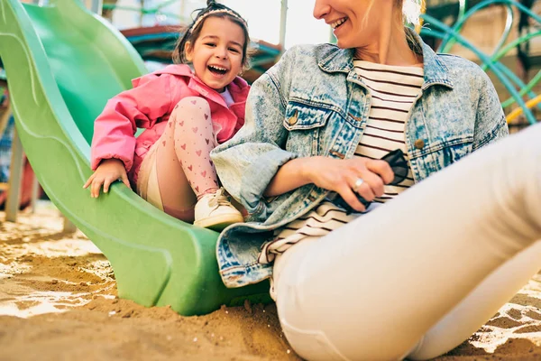 Image extérieure horizontale d'une jolie petite fille souriante et regardant sa belle mère passer du temps ensemble dans une aire de jeux. Jeune femme et fille se sentent heureuses, s'aiment à l'extérieur . — Photo