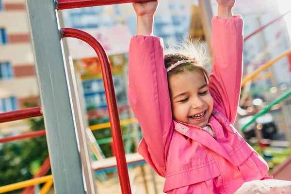 Открытое изображение счастливой маленькой девочки, широко улыбающейся, в розовом наряде, позирующей на детской площадке. Веселый ребенок играет на детской площадке в парке. Счастливое детство, концепция образования . — стоковое фото