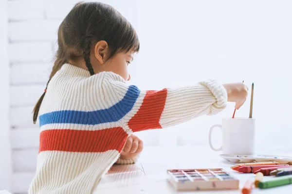 Imagen vista lateral de la linda niña caucásica pintando con pincel sobre papel, sentada en el escritorio blanco en casa. Hermoso dibujo infantil con acuarela. Concepto de personas, infancia y educación — Foto de Stock