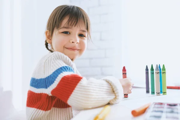 Портрет милой маленькой девочки, улыбающейся и играющей с красочными карандашами. Красивый ребенок рисует с красочными карандашами на бумаге за столом, сидит дома. Человек, детство, образование — стоковое фото