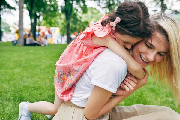 Imagem de perto da criança abraçando a mãe no parque. Bonita menina piggybacks sua jovem mãe atraente sentado na grama verde fora. Amo a emoção. Feliz Dia das Mães. Maternidade e infância — Fotografia de Stock