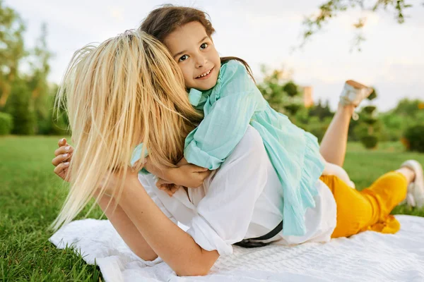 Imagen franca al aire libre de un niño feliz relajándose con su madre, disfrutando del tiempo juntos. Linda niña abraza a su madre sentada en la hierba en el parque. Madre e hija comparten el amor . — Foto de Stock