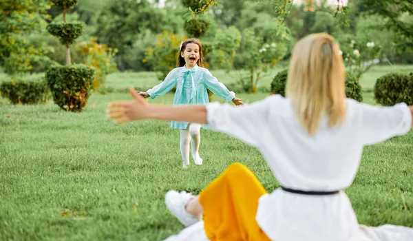 Widok z tyłu szczęśliwego dziecka uśmiechnięty i uruchomiony do jej mama z szerokimi ramionami otwarte w parku, siedząc na zielonej trawie. Ładniutka córka gra z jej młody matka na wolnym powietrzu. Dzień matki. Macierzyństwo, dzieciństwo — Zdjęcie stockowe