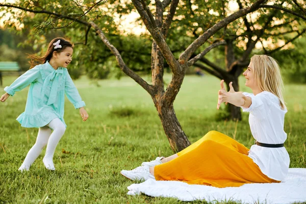 Wizerunek szczęśliwego dziecka biegnącego do jej szczęśliwej mamy z szerokimi ramionami otwartymi w parku, siedząc na zielonej trawie. Ładniutka córka gra z jej młody matka na wolnym powietrzu. Dzień matki. Macierzyństwo i dzieciństwo — Zdjęcie stockowe