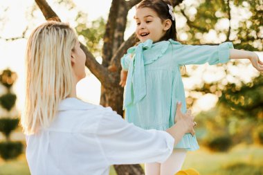 Mutlu kızı gülümseyen ve annesi sarılma, parkta birlikte vakit geçirmek arka görüş görüntüsü. Güzel genç kadın yeşil çimenler üzerinde çocuğuile oynuyor. Anne ve küçük kız sevgipaylaşıyor.