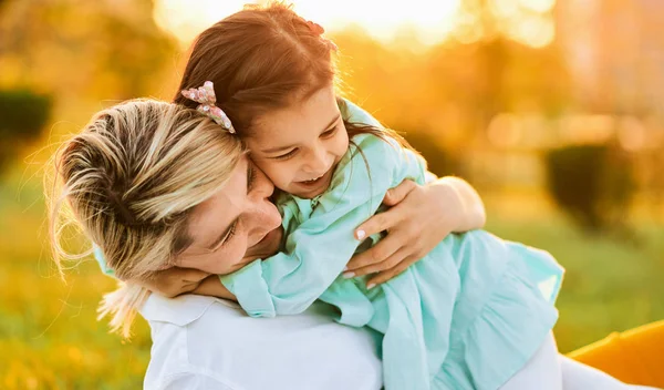 Outdoor portrait of pretty daughter smiling with her mother, spending time together in the park. Beautiful young woman playing with her child on the green grass. Mom and little girl shares love. — Stock Photo, Image
