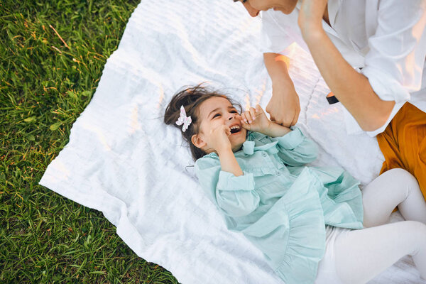 View from above of cute litttle girl playing and having fun with her mother on white blanket at the green grass. Mom and little girl shares love. Mother and daughter spending time together in the park