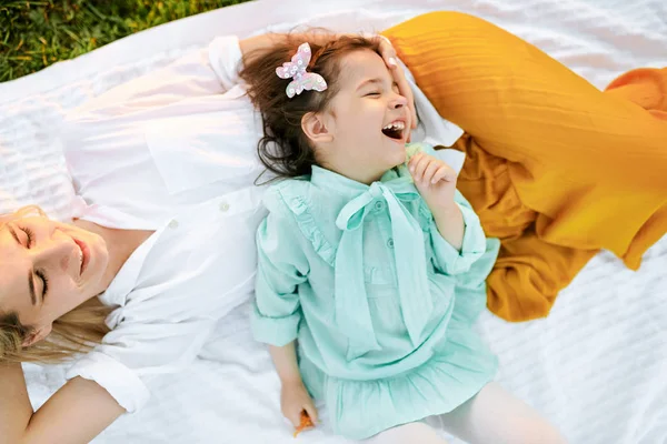 Linda niña jugando y divirtiéndose con su madre en manta blanca en la hierba verde. Mamá y la niña comparten el amor. Madre e hija sonriendo y pasando tiempo juntas en el parque — Foto de Stock