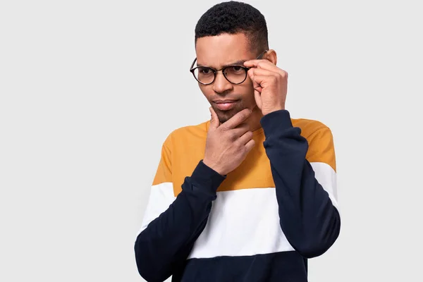 Portrait en studio rapproché de jeune homme afro-américain portant et regardant à travers des lunettes rondes à la mode. Homme d'affaires sérieux portant un pull coloré posant sur un mur de studio blanc. Concept de personnes — Photo