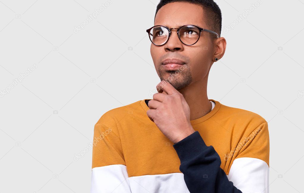 Closeup portrait of young pensive African American man wearing and looking through round trendy spectacles. Serious businessman wearing colorful sweater posing on white studio wall. People concept