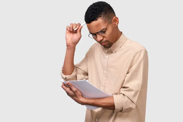 Портрет серьезного молодого африканского студента в бежевой рубашке, анализирующего сообщения, позирующие на белом фоне студии. Мужчина-предприниматель проверяет бумажные бухгалтерские документы. Бизнес, человек — стоковое фото