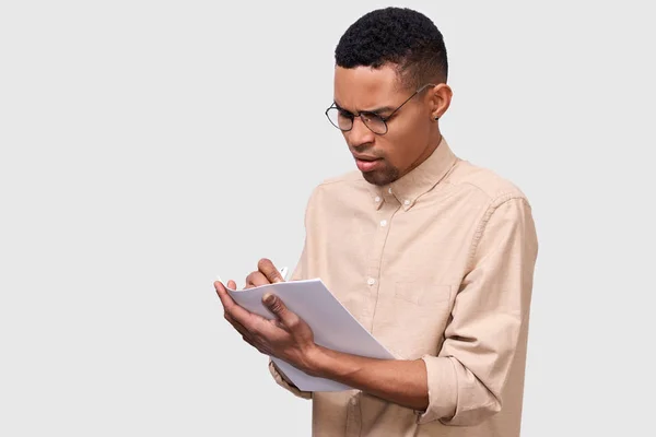 カジュアルなシャツを着た真面目な若いビジネスマンの屋内肖像画は、白いスタジオの背景にポーズをとって財務報告を分析します。アフリカ系アメリカ人男性起業家が紙会計書類をチェック. — ストック写真