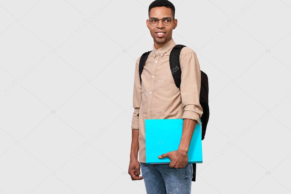 Indoor shot of cheerful male student in casual outfit with blue folder in hand, with backpack smiling and looking to the camera. Young African American businessman holding folder with documents 