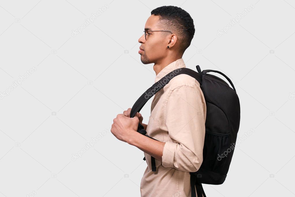 Side view image of young Afro man student with backpack standing over white wall. Young businessman wearing casual beige shirt and round eyewear looking at blank copy space for your promotional text.