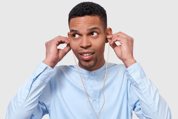 Portrait d'un homme à la peau foncée attirant mis des écouteurs pour écouter la musique. Plan intérieur de jeune homme afro-américain porte chemise bleue, regardant loin. Personnes, technologie et concept de musique — Photo