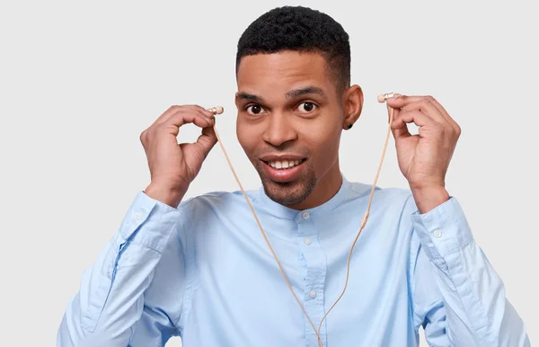 Portrait en studio d'un homme heureux à la peau foncée touchant des écouteurs, écoute la musique. Photo intérieure de jeune homme afro-américain portant une chemise bleue, regardant vers la caméra. Concept de personnes — Photo