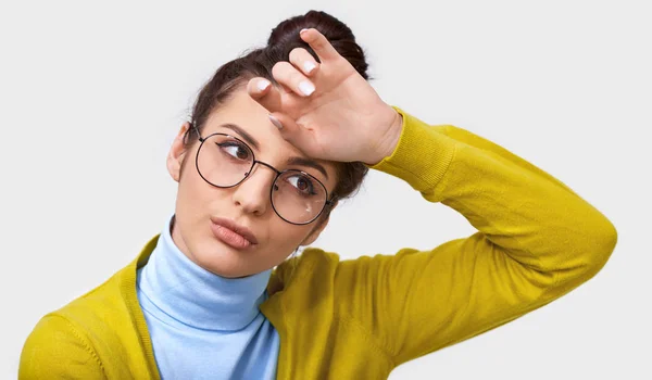 Retrato de cerca de una mujer joven y bonita cansada con moño de pelo, con un atuendo casual con gafas redondas de moda, mantiene la mano en la frente, mientras está de pie sobre la pared blanca. Concepto de personas y emociones —  Fotos de Stock