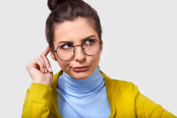 Portrait d'une jeune femme incertaine coiffée d'un chignon, portant une tenue décontractée et des lunettes rondes transparentes, regardant d'un côté avec la main sur la tête, isolée sur fond de studio blanc . — Photo