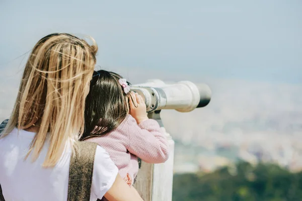 Vista posteriore della bella madre che tiene sua figlia guardando una splendida vista sulla città attraverso un binocolo a moneta. Mamma e bambina viaggiano insieme durante il viaggio. Concetto di viaggio — Foto Stock