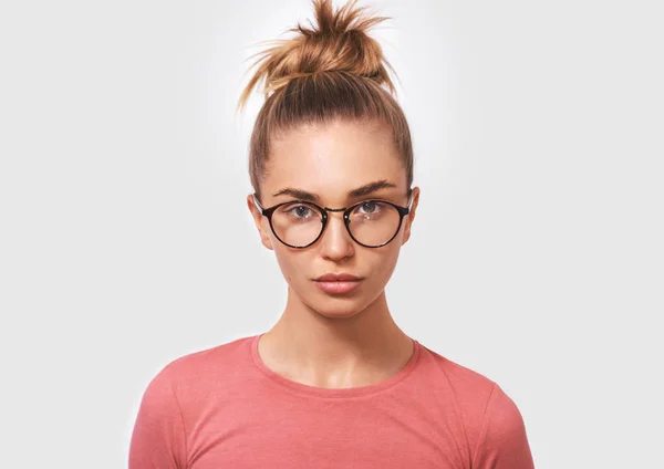 Portrait of attractive blonde young woman wearing round transparent spectacles and casual pink blouse, looks seriously directly into camera, poses against white studio background. People and emotions — Stock Photo, Image