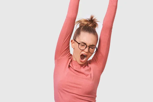 Image of tired Caucasian young woman yawning, wearing pink blouse and round transparent eyewear, posing over white studio wall. Sleepy female with hair bun, feel sleepy. People and emotions concept