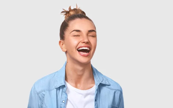 Happy female student with bun hairstyle smiling positively and being in great mood while standing over white studio background. Beautiful young woman wearing blue denim shirt smiling broadly. People — Stock Photo, Image