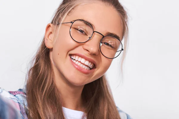 Primer plano retrato feliz hermosa mujer joven positiva vestida con traje casual, usando gafas redondas transparentes con agradable sonrisa ampliamente, mirando a la cámara y posando sobre la pared del estudio blanco —  Fotos de Stock